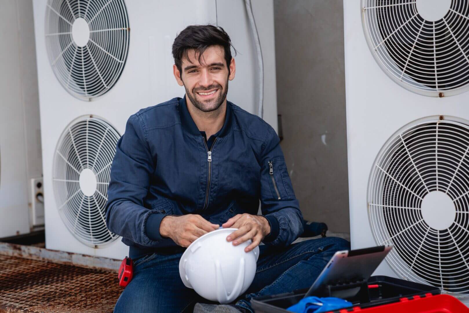 Smiling man in workwear holding a white hard hat indoors.
