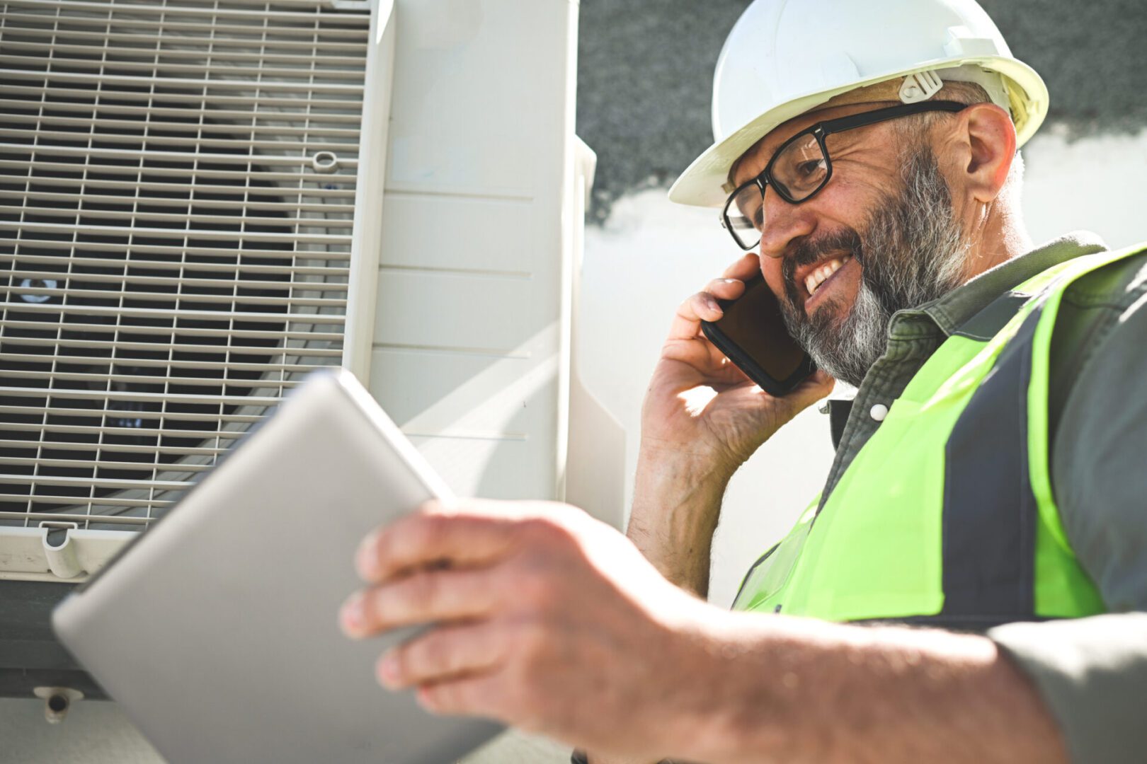 Technician in safety gear inspecting HVAC system while talking on phone.